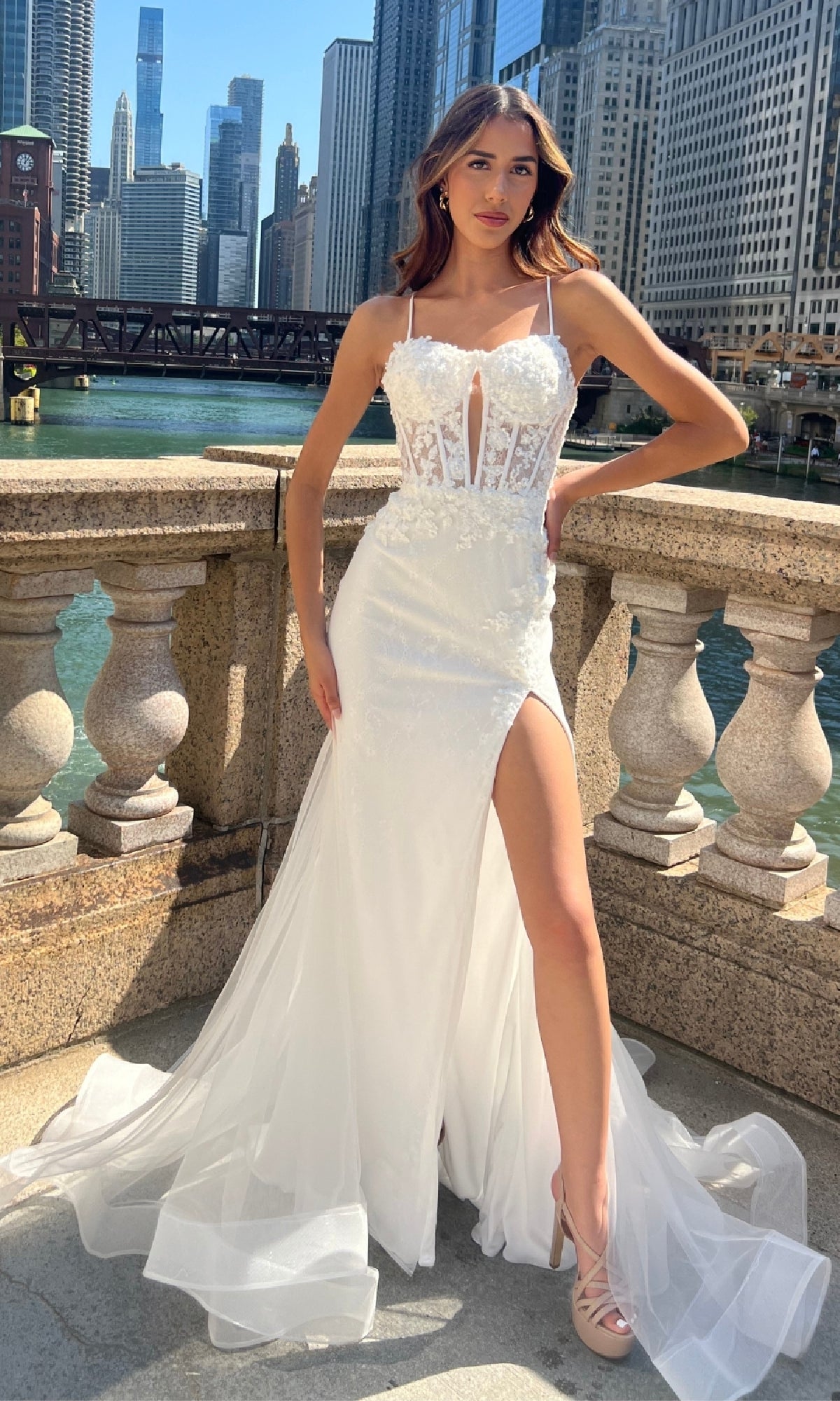 A woman in a white, sleeveless lace Long Mermaid Prom Dress, Amelia Couture 7064, with a high slit, stands on a stone balcony by the river, radiating elegance in the city.